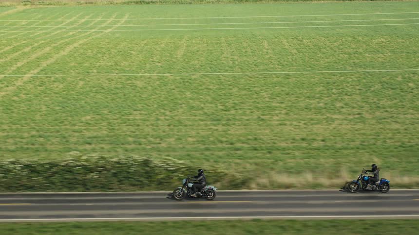 Two motorcyclists ride on a singlelane road surrounded by vast green fields