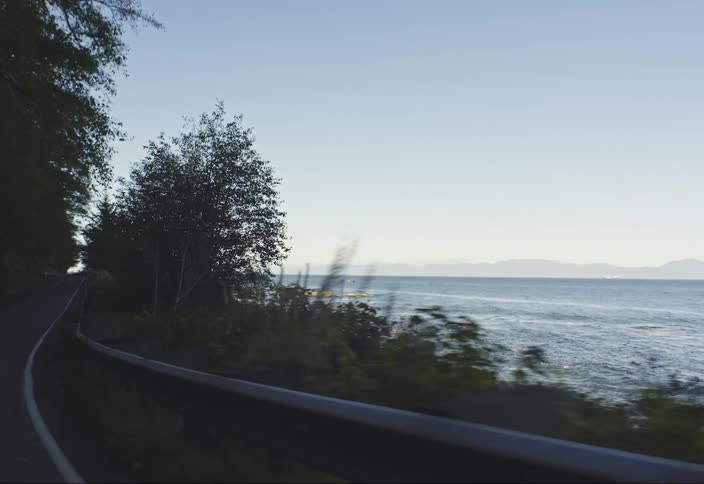 A vehicles perspective the winding asphalt road bordered by a metal guardrail curves sharply to the left along a rocky uneven shoreline where waves gently lap against the stones with a backdrop of a calm vast ocean stretching to the horizon