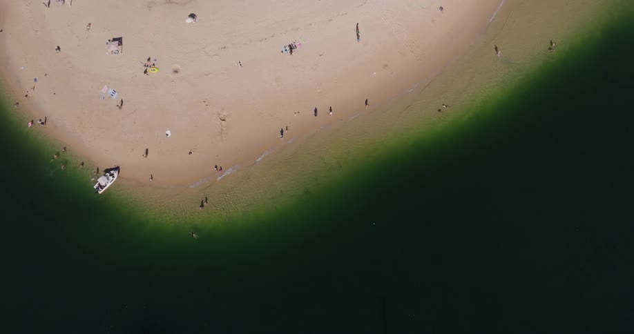 An aerial shot shows a mostly deserted beach with a few scattered people and a single boat with a large shaded area in the foreground