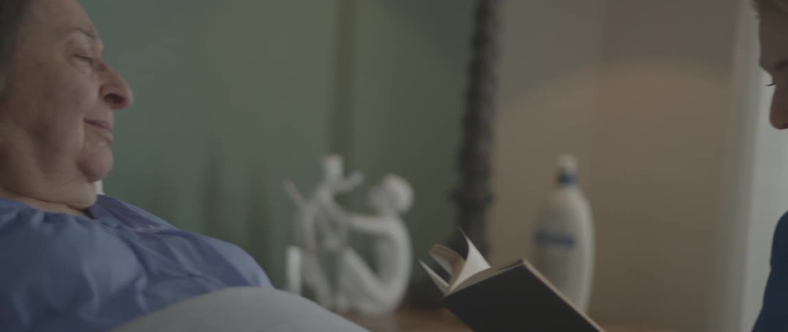 A woman smiles and reads from a book to an elderly woman
