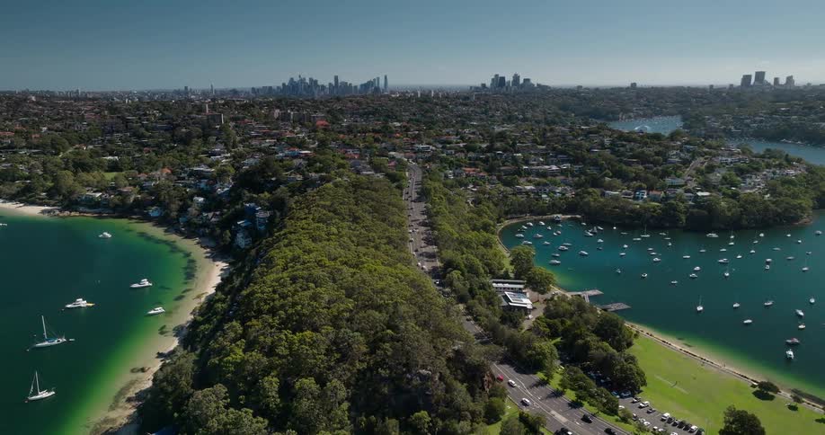 Aerial view shows a road surrounded by dense vegetation between two bays with anchored boats with a city of skyscrapers in the background