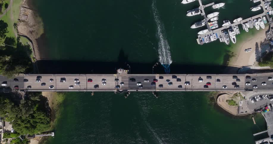 A gray bridge crowded with cars spans a dark green river or inlet connecting two landmasses with marinas and docked boats on either side under a clear sky