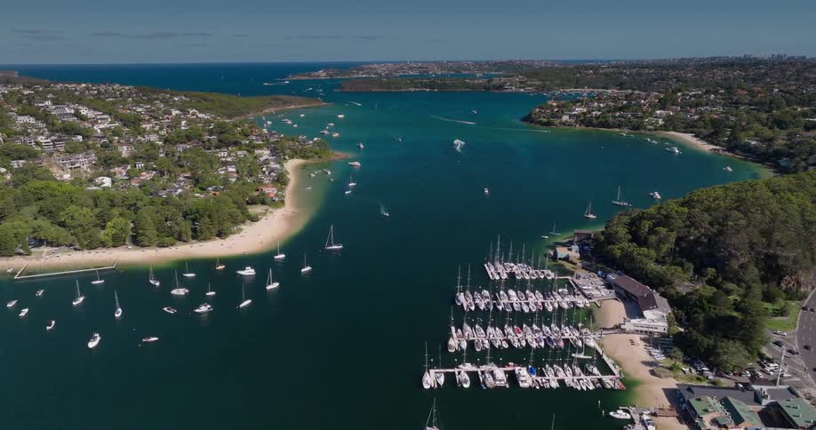 Aerial view of a wide deep blue bay filled with numerous white sailboats and yachts some moored closely together others sailing further out with a busy highway running along the densely populated shoreline bordering the water which also features a sm