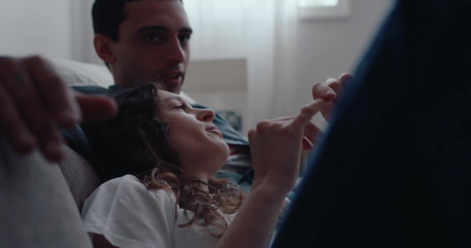 A young woman reclines against a young man on a couch indoors and she is gesturing with her hands as if speaking passionately