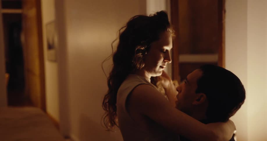 A dimly lit indoor scene shows a man holding a woman in his arms as they look at each other closely creating an intimate and emotional moment