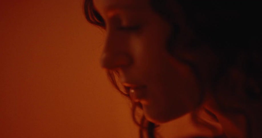 A woman with curly hair shows her in warm dim lighting speaking softly with a calm and introspective expression on her face