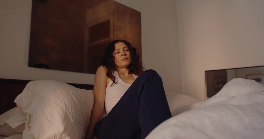 A woman with curly hair wearing a white tank top and black pants gets out of bed in her bedroom