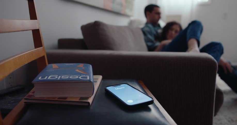 A woman with curly hair reclining on her boyfriends lap on a couch rises to answer her ringing iphone