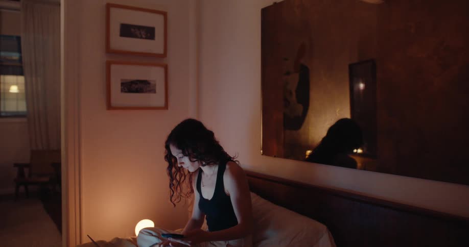 A woman sits on her bed opens her laptop and begins scrolling on her smartphone