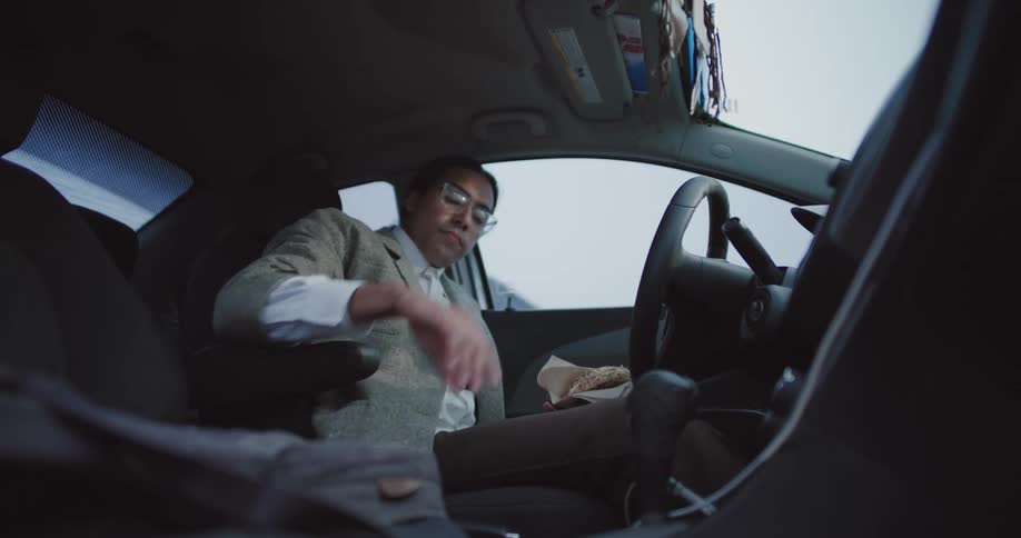 A person sits in the drivers seat placing a water bottle in the cup holder while holding a sandwich