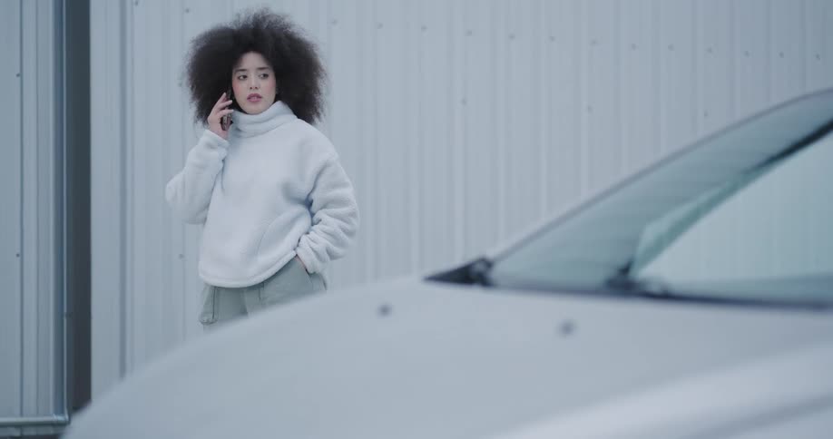 A woman with long hair and wearing white clothes and is talking on the cell phone while walking forward on a street and then she stops and looks at a car that is parked and makes a denial sign and continues