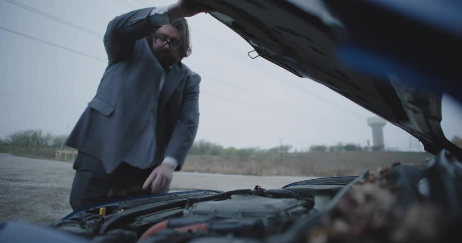 A man in a suit with disheveled hair and glasses is standing on the side of a rural road leaning over an open car hood with a concerned expression