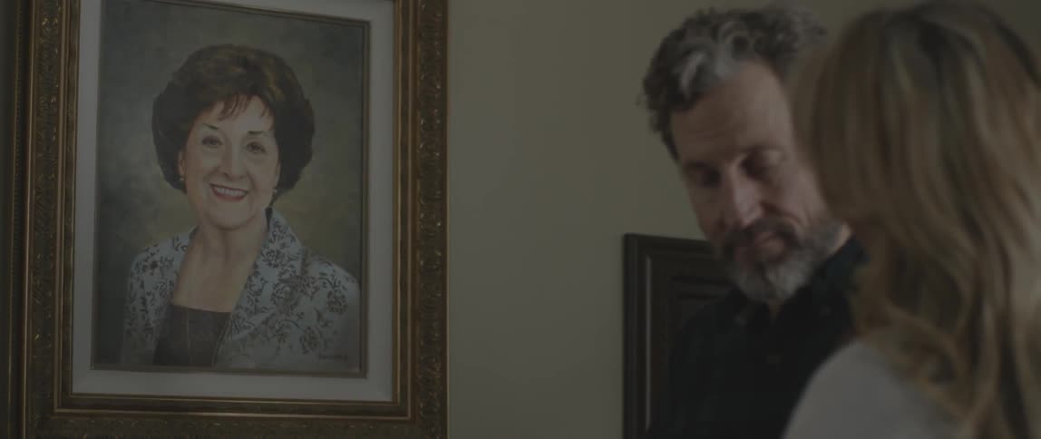 Two people stand in a home and look at a portrait on the wall
