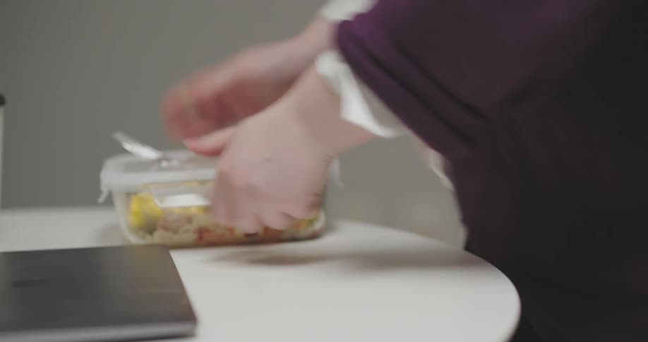 The face of a blonde woman with glasses who is taking a lunch box of food from a microwave and then sitting down