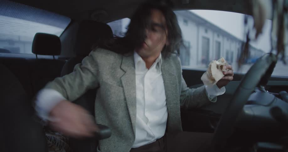 A man eating a sandwich in a car removes his ponytail and starts headbanging to music