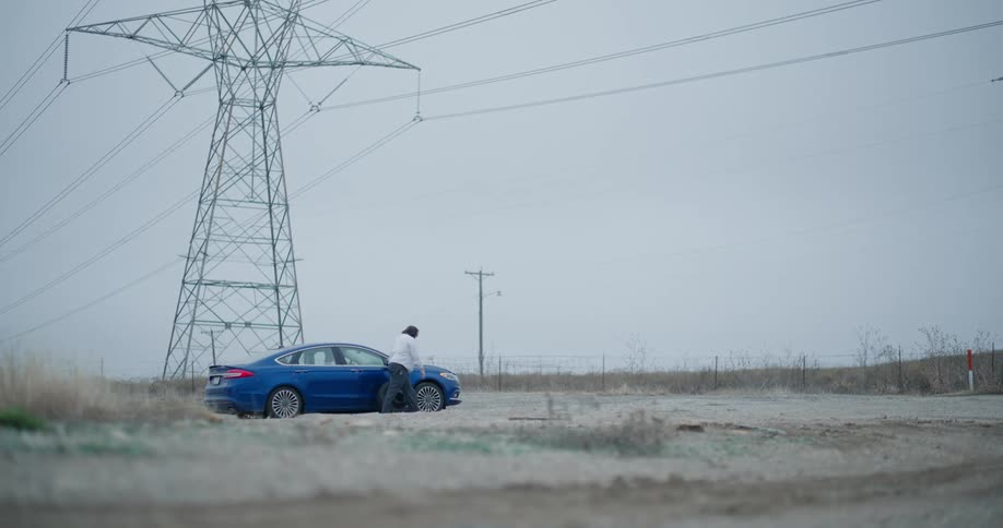 Man removing a tire from the back seat of a car that is stopped on a deserted road