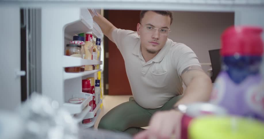 A person is kneeling in front of an open refrigerator looking inside with various food items visible on the refrigerator door shelves