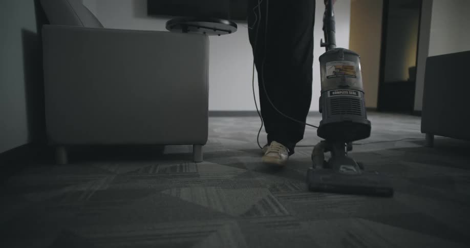 A woman wearing black pants and dirty white sneakers vacuums a gray and white patterned carpet in a dimly lit room as seen from a low angle