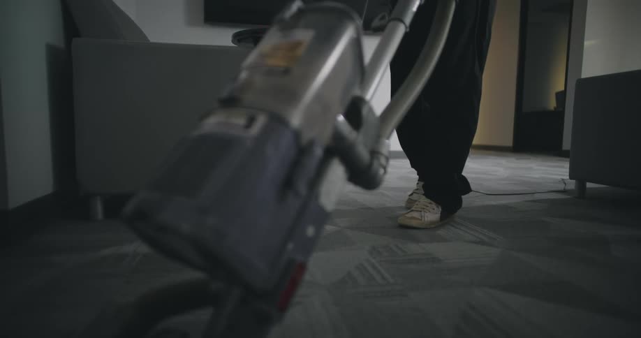 A person is vacuuming the carpeted floor in an officelike room with a gray chair and a wallmounted television in the background
