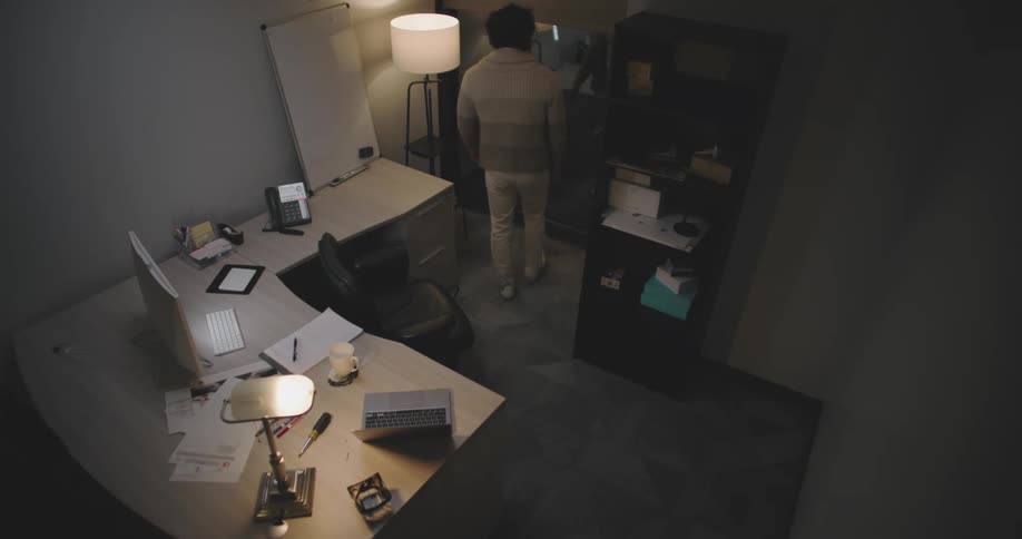 A man in a grey wool jacket and white pants paces around a home office passing a desk with a macbook and imac as he appears to wait
