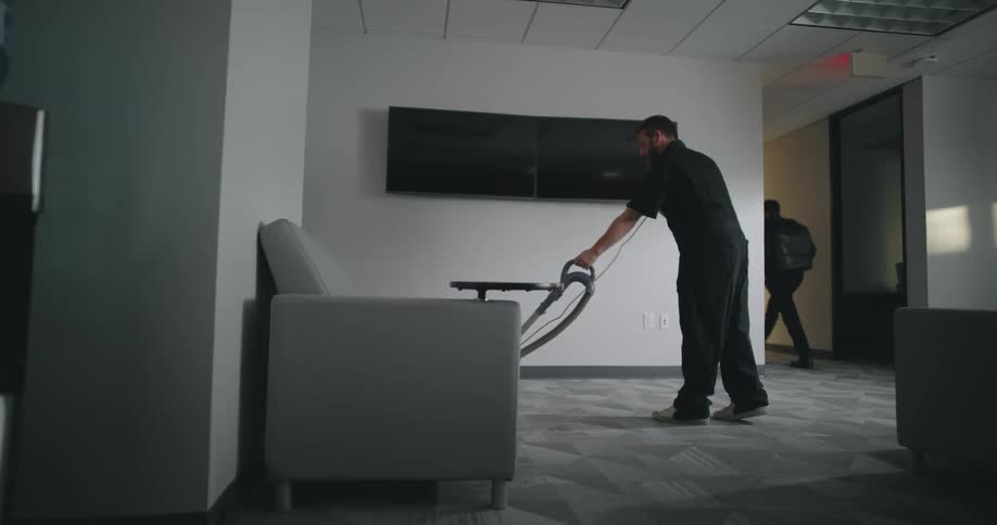 A person is vacuuming the carpeted floor in an officelike room with a water cooler a gray chair a trash can and a wall mounted television in the background