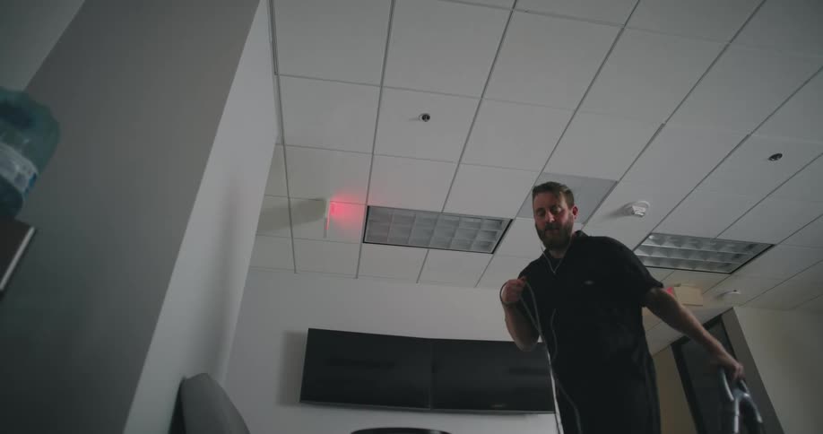 A man in a black shirt vacuuming a room with a modern officelike setting featuring a drop ceiling a mounted television and a couch