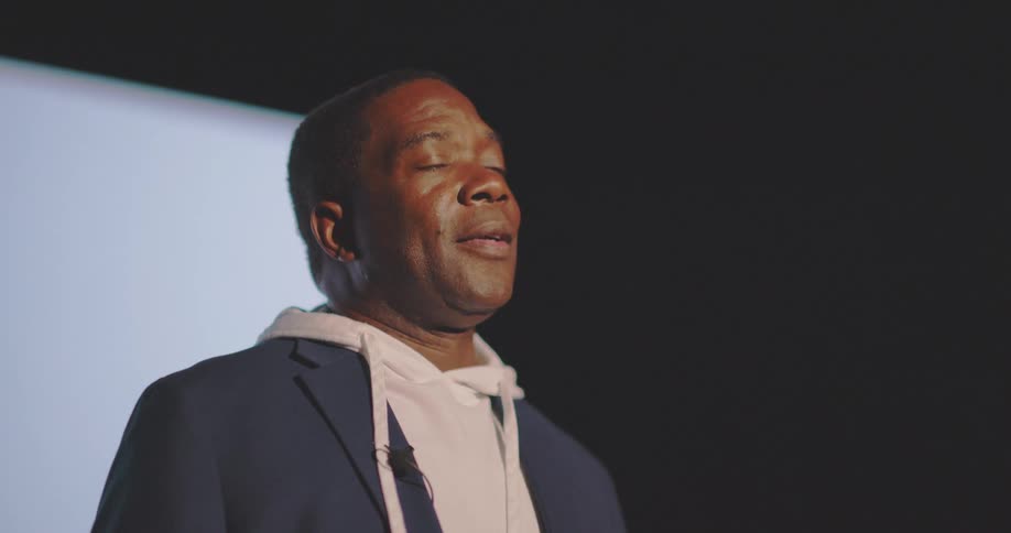 A man wearing a hoodie under a blazer is midpresentation with an intense expression as camera flashlights illuminate him against a bright screen backdrop