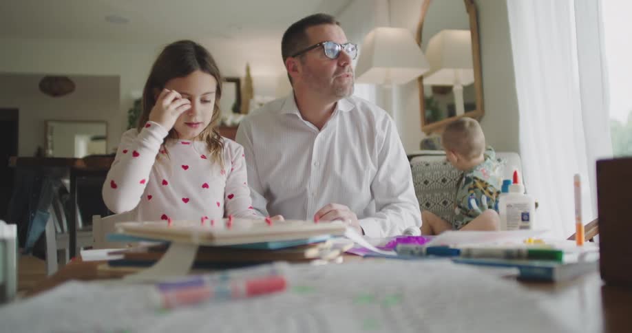 Man wearing glasses and a white shirt interacts with three children who are gathered at a table full of art supplies in a room lit by natural light