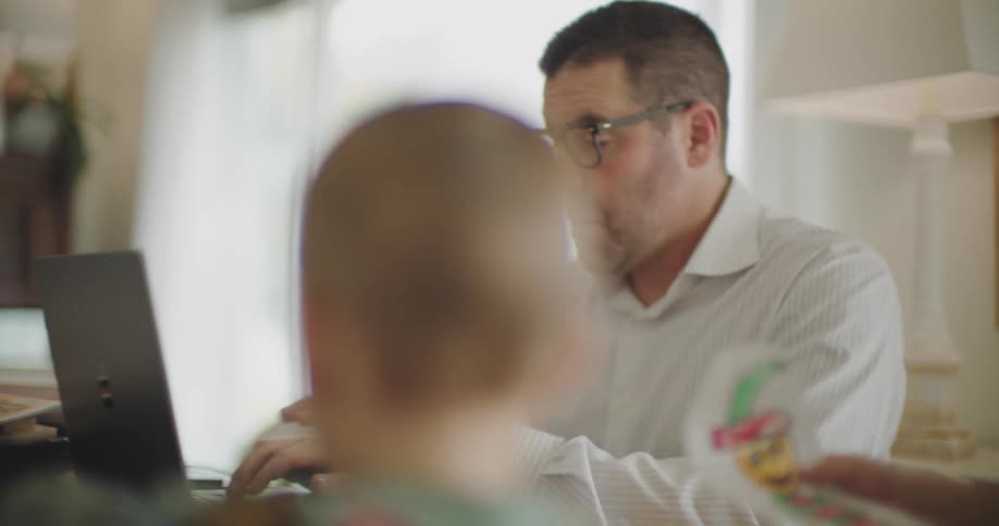 A father working from home in a white shirt sits with his children around trying to work on a laptop in front of them