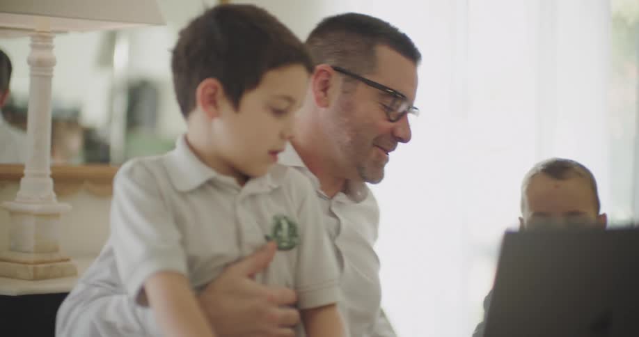 A father working from home in a white shirt sits with his children trying to work on a laptop in front of them