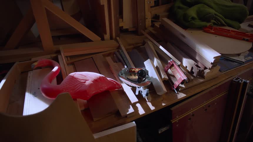 A pink plastic flamingo stands out amidst a collection of wooden planks and various objects in a dimly lit cluttered space