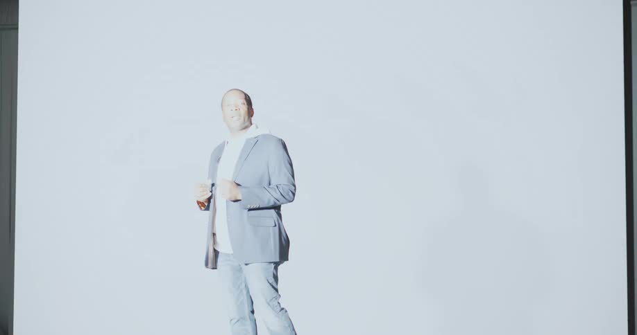 Speaker entering a stage and receiving several flashes of photography against a screen in the background