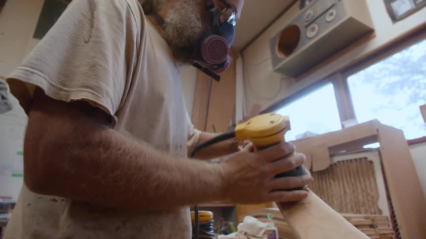 A person is working in a woodworking shop using a power sander on a piece of wood surrounded by various tools and equipment