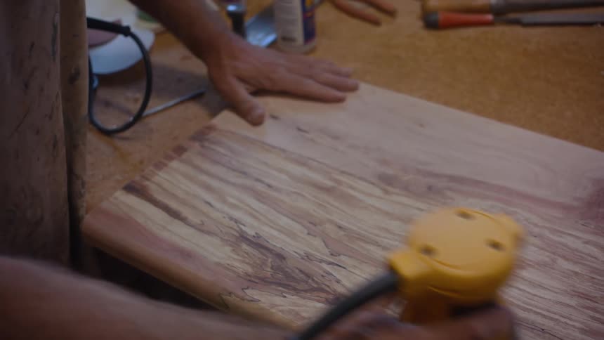 Man sands a wooden board with a yellow electric sander on a woodworking bench