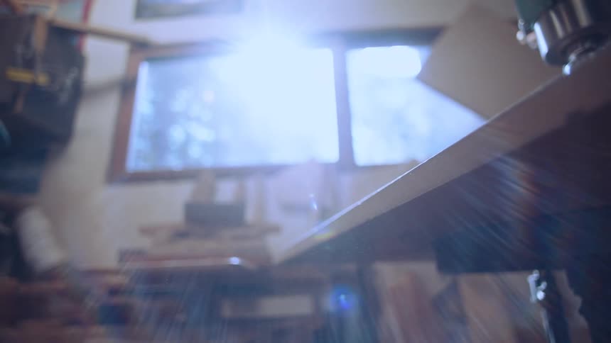 A sunlit workshop interior is seen through a window and a partially obscured wooden table