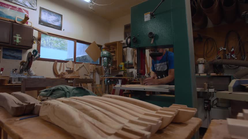 A person works at a bandsaw in a cluttered workshop with several lightcolored wooden pieces arranged in the foreground