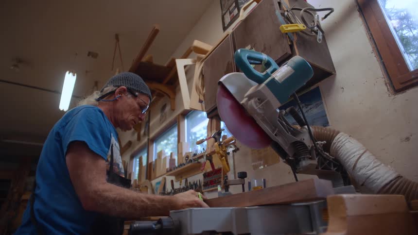 A person is operating a miter saw in a woodworking shop filled with various pieces of wood and tools