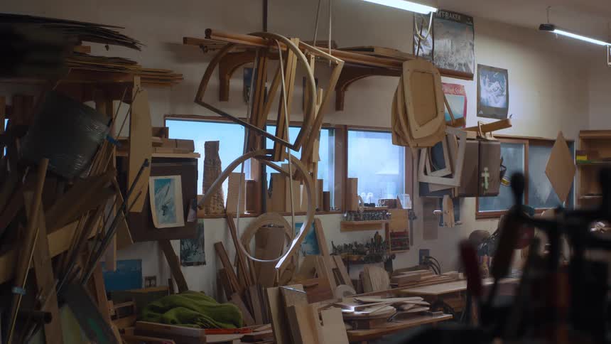 A cluttered woodworking shop features wooden frames hanging from the ceiling surrounded by paintings sculptures and assorted materials