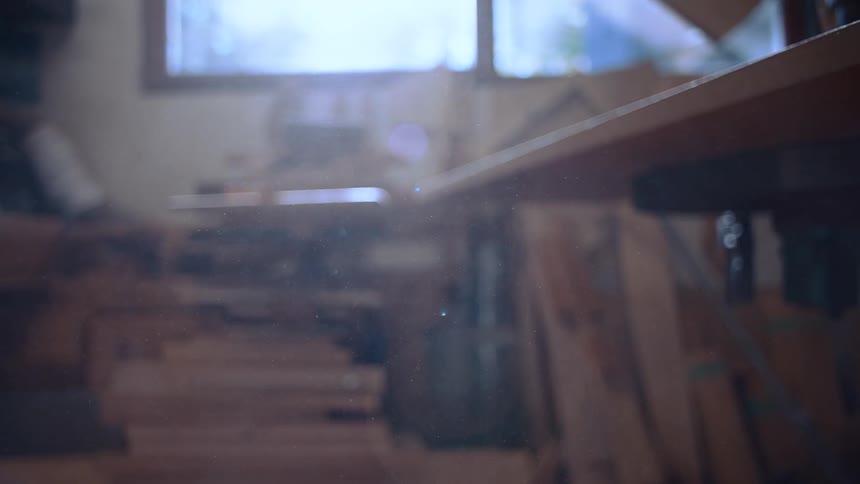 A sunlit workshop interior is seen through a window and a partially obscured wooden plank