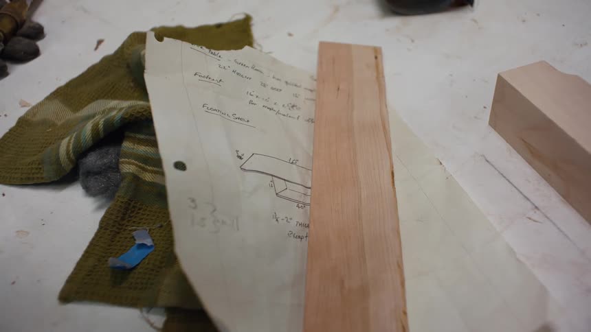 A wooden plank rests on a paper with handwritten notes and a sketch next to a folded green cloth