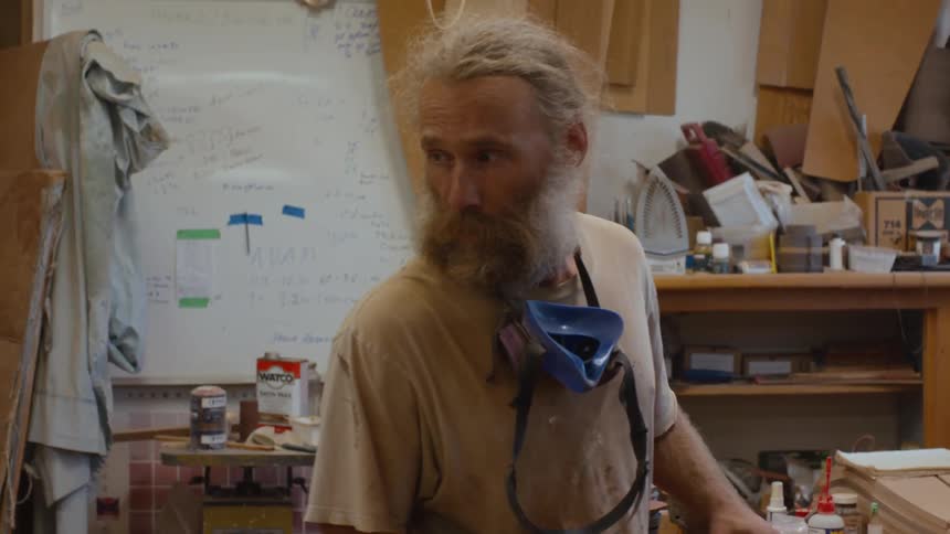A bearded male in a workshop handling a piece of wood and talking