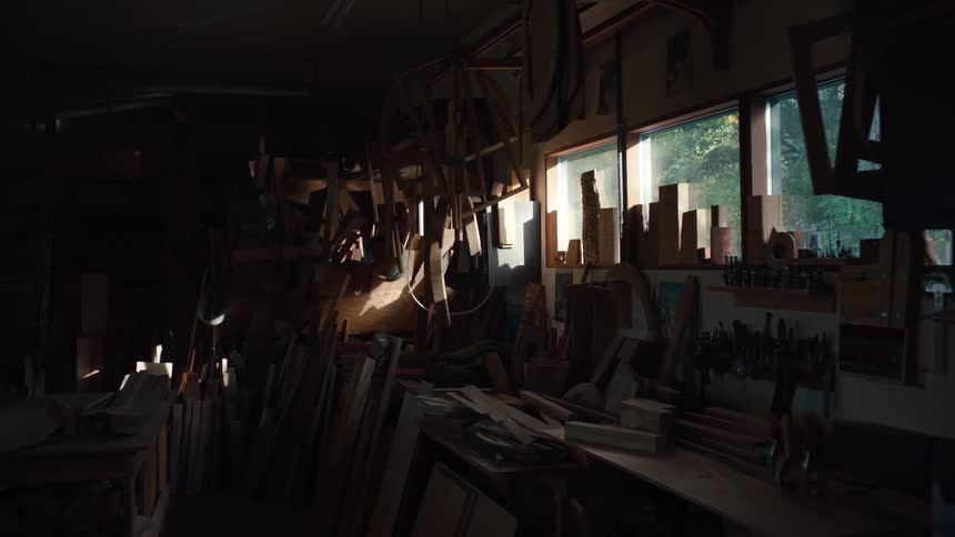 Sunlight streams through a window into a cluttered workshop illuminating hanging tools and various wooden scraps