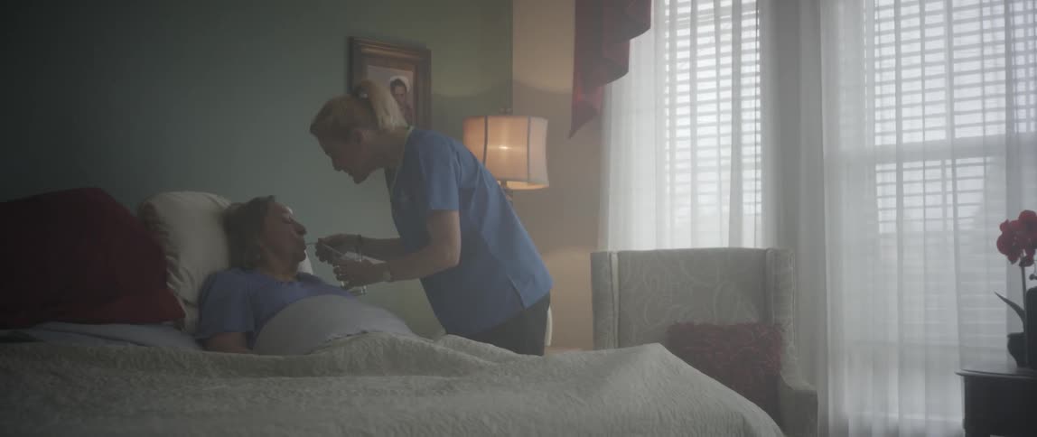 A woman brings a glass of water to an elderly woman laying in bed