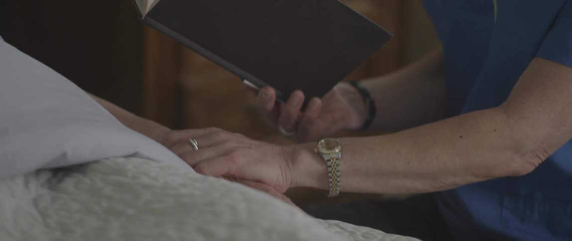 Elderly woman reading to someone lying in a bed and then reaching out and holding their hand