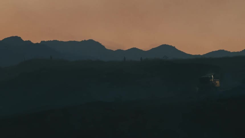 Silhouetted people walk atop hills under a dusky sky with a small indistinct vehicle visible on one dustcovered slope