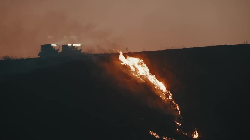 A dramatic scene of a fire spreading down a hillside at dusk with two firefighting bulldozers in the background against a smoky orange sky