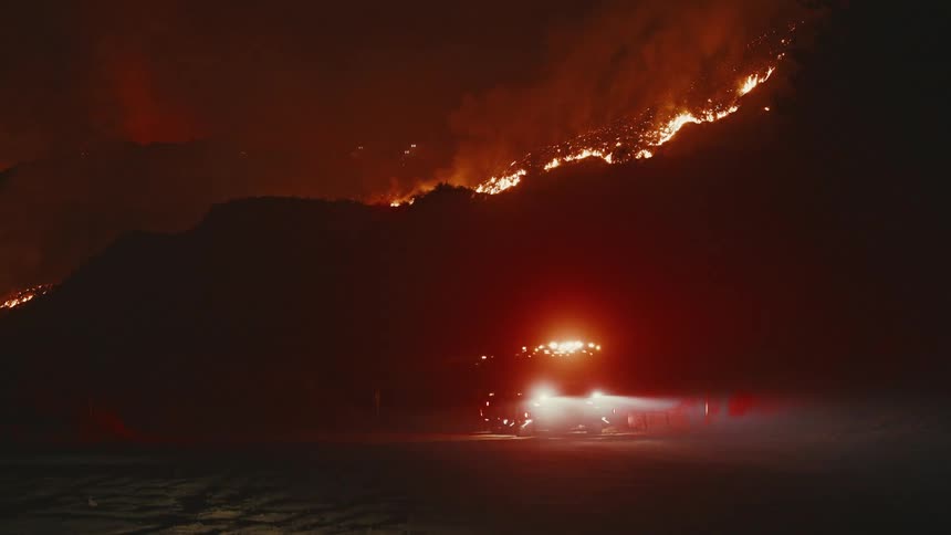 A wildfire blazes across a rural hill illuminating thick smoke as a fire truck with flashing lights is parked near the base of a hill on a dirt road