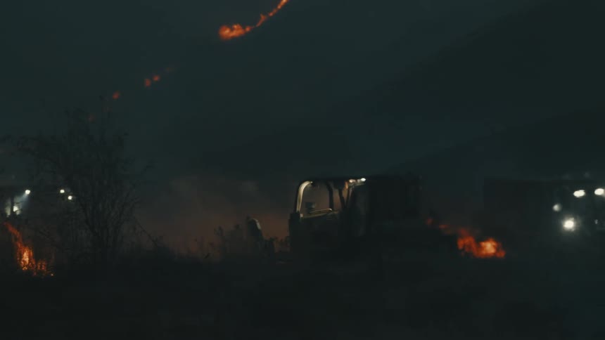 At night a tractor plows the field with its headlights on helping control the hillside fire as flames glow above