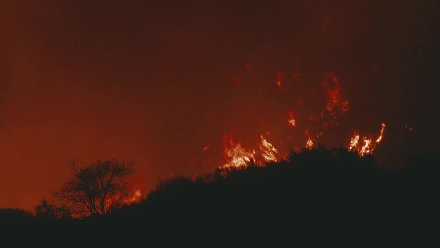 A dark silhouette of a treelined ridge is engulfed by bright orange flames against a dark red smokefilled sky