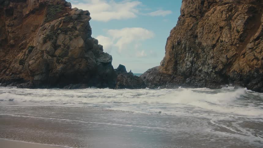 A wave crashes between two towering rock formations revealing the vast ocean beyond as waves roll onto the sandy shore in the foreground